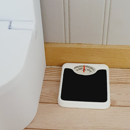 Closer front view of miniature bathroom scales showing the white case, black foot pad and red dial pointer