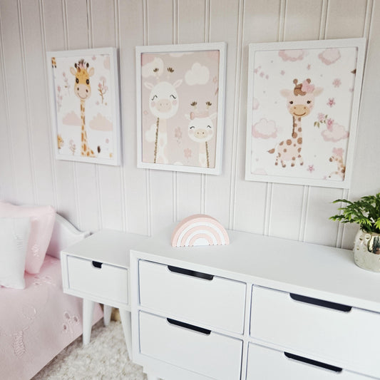 May include: Three framed giraffe prints in a nursery. The white-framed pictures show giraffe illustrations on pink and beige backgrounds. A white dresser with drawers and a wooden rainbow decoration are in the foreground. The room has white panelled walls.
