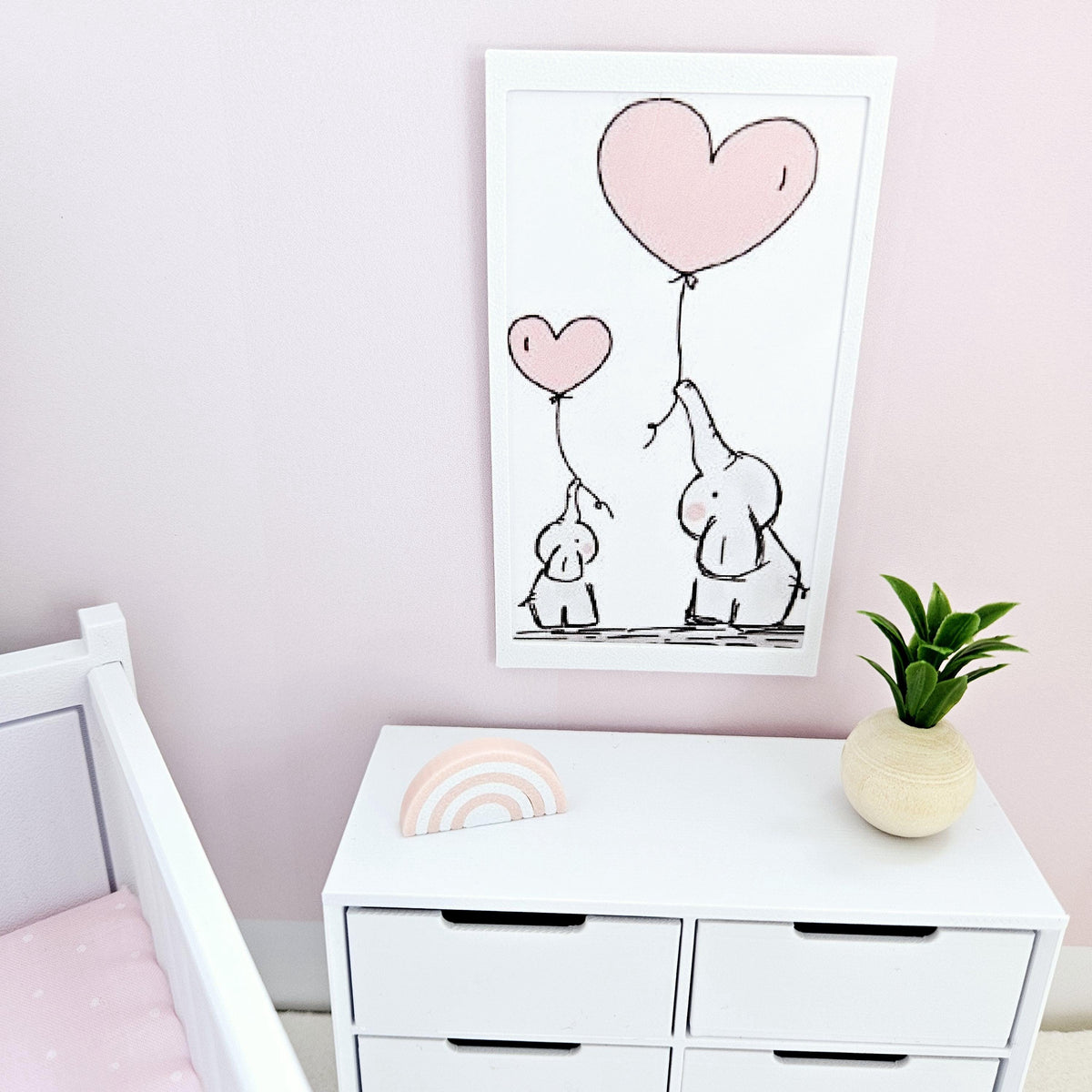 May include: A framed art print showing two cartoon elephants with pink heart-shaped balloons. The artwork is in a white frame and hangs on a pink wall above a white chest of drawers. A small rainbow and a plant in a vase are on the drawers.