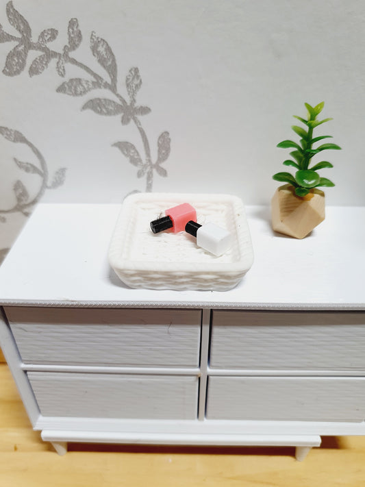 May include: Miniature pink and white nail polish bottles sit in a small, white, textured dish. A small, green plant in a geometric beige pot is on a white dresser. The background has a subtle floral design.
