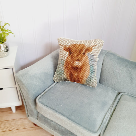 May include: A light blue velvet sofa with a square cushion featuring a brown Highland cow with white markings. The cushion has a white border.