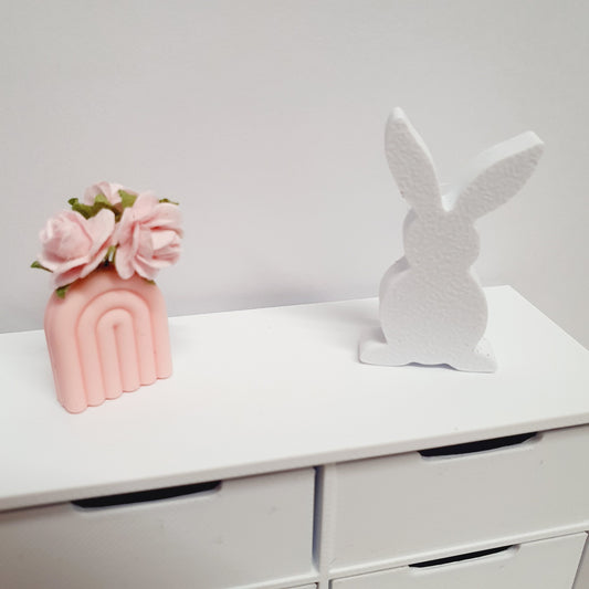 May include: A white wooden bunny figurine and a pink ceramic rainbow vase with pink flowers on a white dresser with drawers.