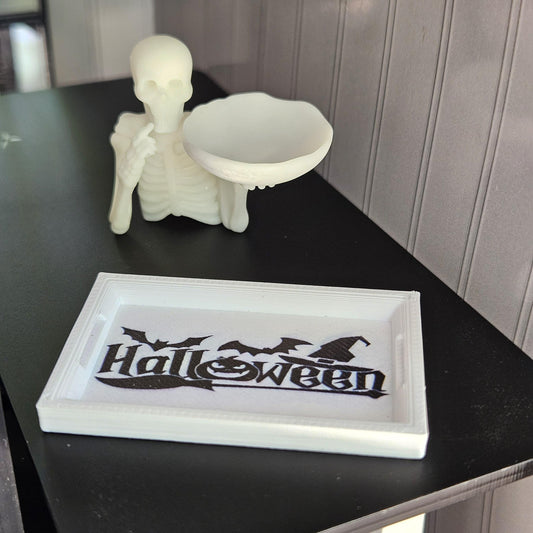 May include: A white Halloween tray with the word "Halloween" in black script, bats, and a witch hat. A glowing skeleton figure holds a white bowl. The items are on a black surface.