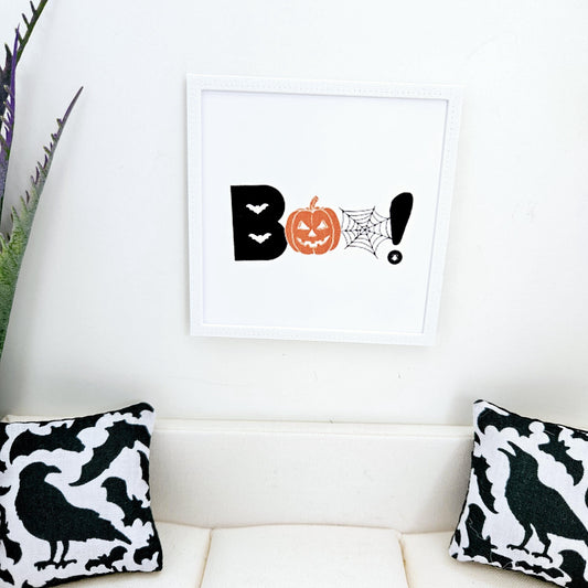 May include: A white framed print with the word "BOO!" in black letters, a jack-o'-lantern, a spider web, and an exclamation point. The print is hanging on a white wall above a white sofa with black and white cushions.