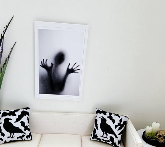 May include: A black and white framed print of a shadowy figure with outstretched arms, appearing to be trapped behind a frosted glass surface. The print is hanging on a white wall above a white sofa with black and white crow patterned throw cushions.