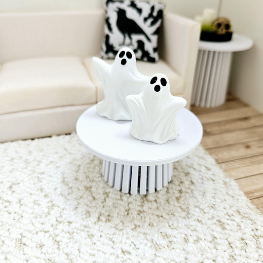May include: Two white ceramic ghost figurines with black eyes on a white round table with a white fluffy rug underneath. The table has a white base with a fluted design.