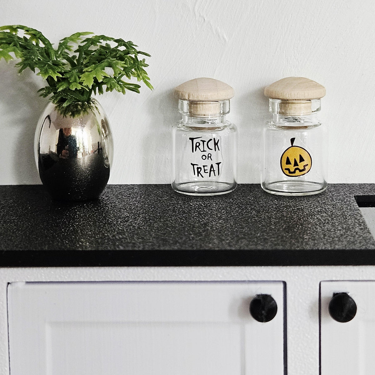 May include: Two small glass jars with wooden lids, one with "Trick or Treat" text and the other with a jack-o'-lantern graphic. A silver vase with green foliage is on the left. The jars sit on a black surface, with white cabinets below.