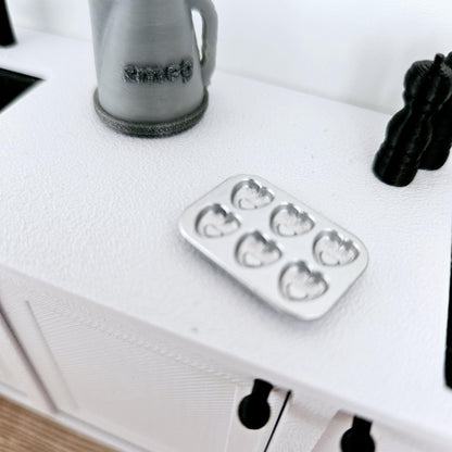 May include: A silver heart-shaped muffin tin with six cavities. The tin is on a white work surface.