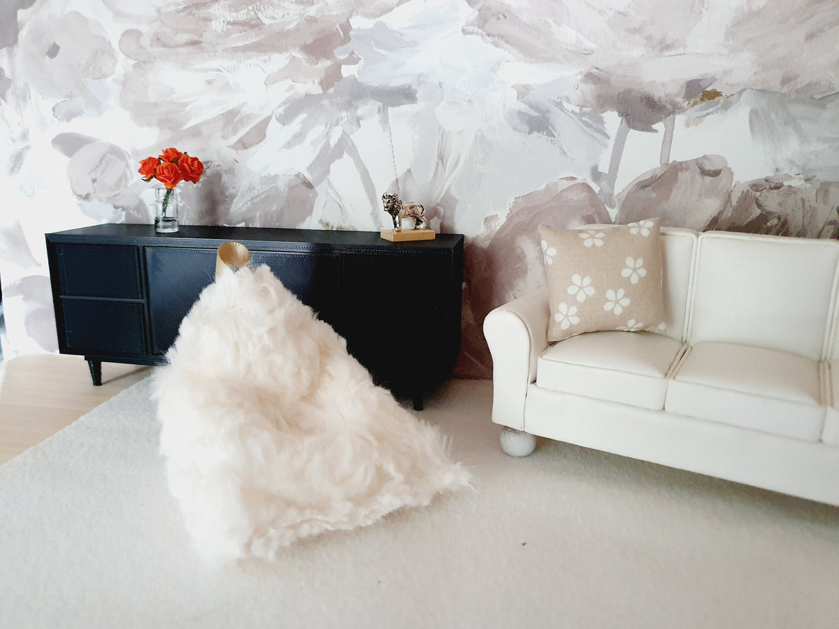 May include: A white faux fur bean bag chair sits on a white rug in a living room. A white sofa with a beige cushion featuring white flowers is in the background. A black cabinet with a small metal sculpture of a bull sits on top of the cabinet.