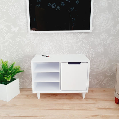 May include: A miniature white entertainment unit with a cabinet door and two open shelves. The unit has a minimalist design with tapered legs. A small fern in a white pot sits to the left. A TV is mounted above the unit.