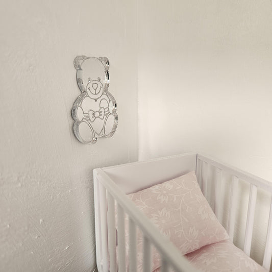 May include: A clear acrylic teddy bear wall decoration is fixed to a white wall. The bear has a bow tie and is holding a heart. Below is a white cot with a pink floral pillow and bedding.