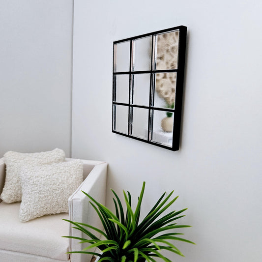 May include: A black framed mirror with nine square panes of glass. The mirror is hanging on a white wall above a white sofa with two white cushions. A green plant is in the foreground.