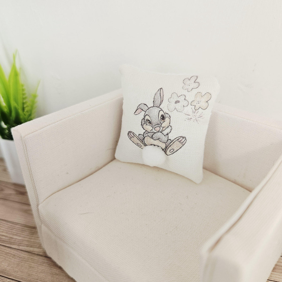 May include: A small, square decorative cushion featuring an embroidered cartoon rabbit with a fluffy white tail. The rabbit is grey and white, and the cushion is on a cream-coloured armchair. The cushion also has embroidered flowers.