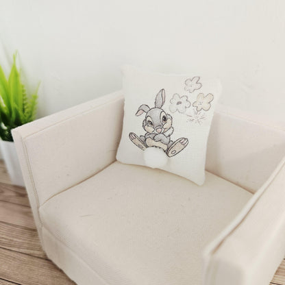 May include: A small, square decorative cushion featuring an embroidered cartoon rabbit with a fluffy white tail. The rabbit is grey and white, and the cushion is on a cream-coloured armchair. The cushion also has embroidered flowers.