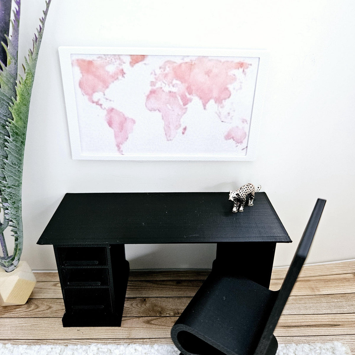 May include: A black desk with a white frame around a pink watercolour world map hanging above it. A small black chair and a leopard figurine are on the desk.