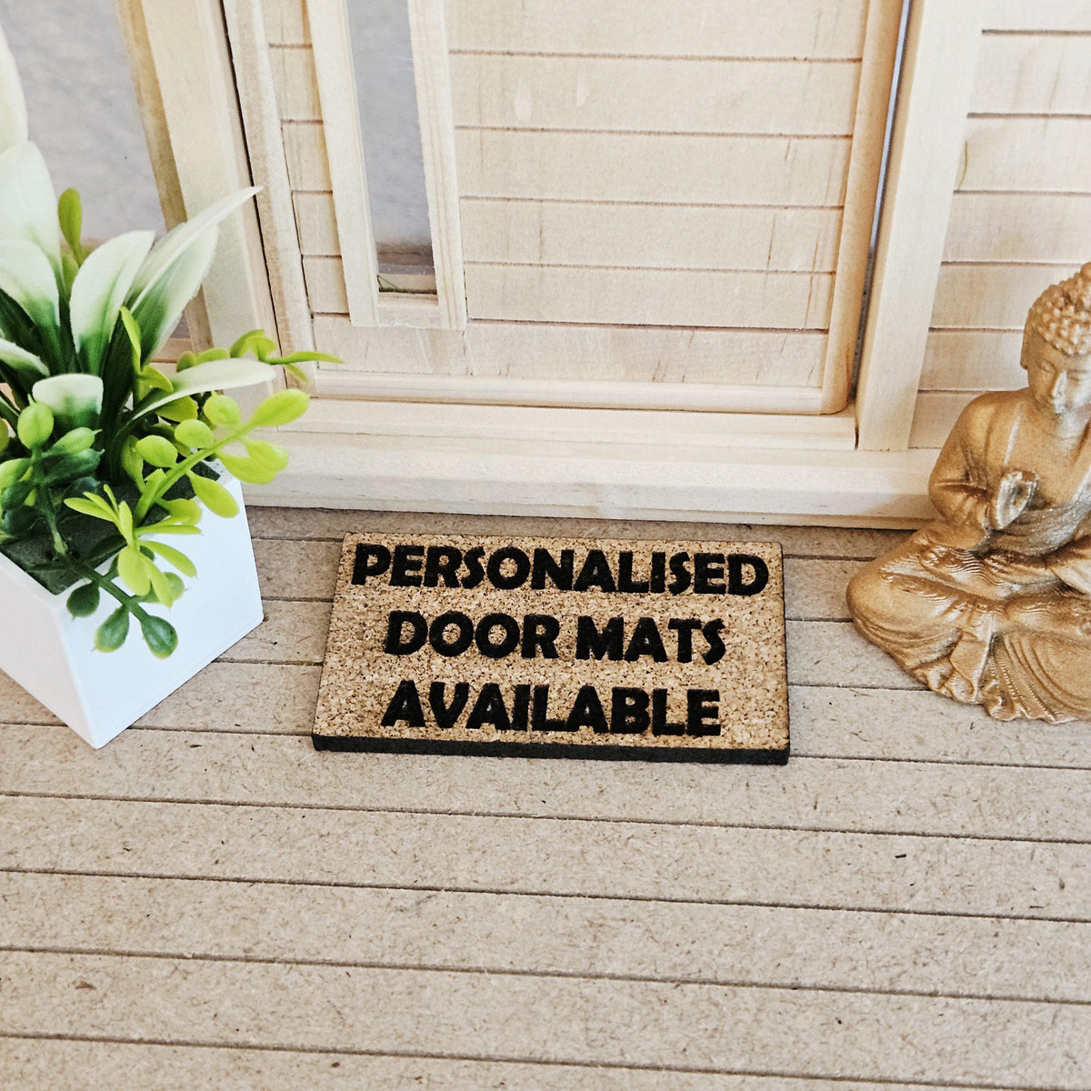 May include: A rectangular doormat with the text "PERSONALISED DOOR MATS AVAILABLE" in black lettering. The mat is made of a light brown material and is placed on a light brown surface. A white square planter with green plants is to the left, and a gold Buddha statue is to the right.