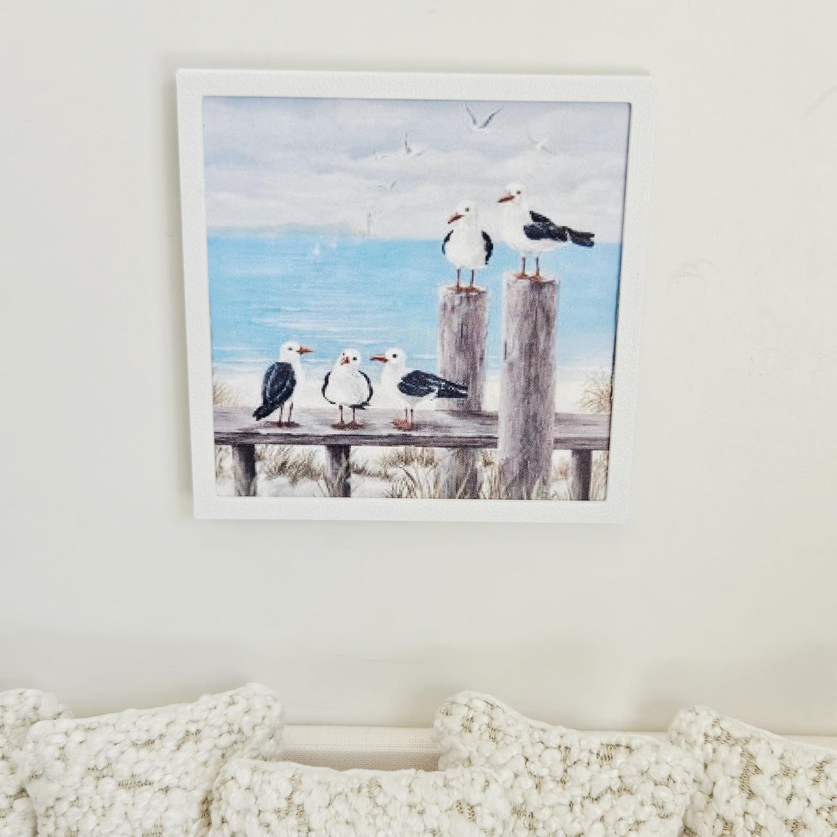 May include: A framed print depicting a coastal scene with five seagulls on wooden posts and a bench. The artwork shows a blue ocean, a cloudy sky, and sailboats in the distance, creating a serene beach theme.