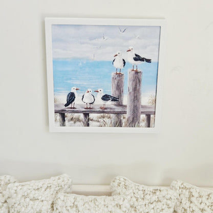May include: A framed print depicting a coastal scene with five seagulls on wooden posts and a bench. The artwork shows a blue ocean, a cloudy sky, and sailboats in the distance, creating a serene beach theme.