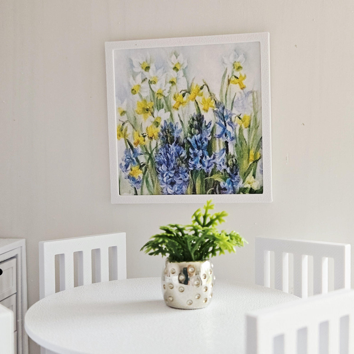 May include: A framed floral print with yellow and white daffodils and blue hyacinths hangs above a white table. A small silver pot with a green plant sits on the table. White chairs surround the table.