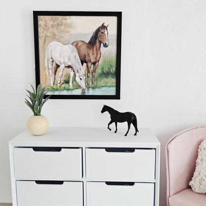 May include: A framed print of two horses, one white and one brown, drinking water, hangs above a white chest of drawers. A black horse figurine stands on the drawers, next to a small plant in a beige vase.