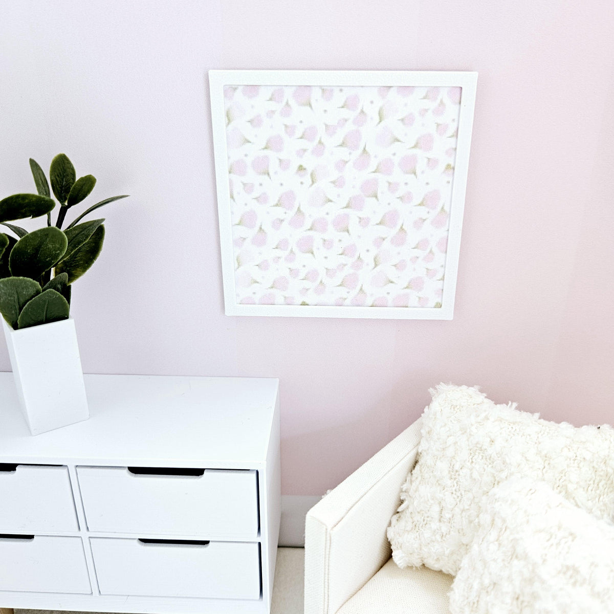 May include: A white framed picture with a pink and white abstract floral pattern on a pale pink wall. The picture is hanging above a white dresser with six drawers. A white armchair with a fluffy white cushion is in the corner of the room.