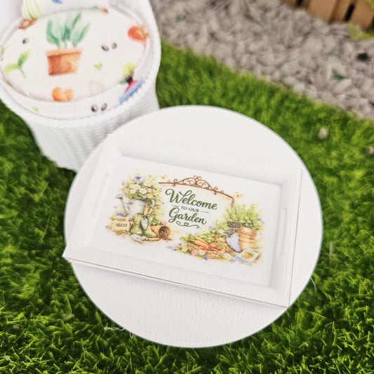 May include: A miniature white framed sign with the text "Welcome to our Garden" and a watercolour illustration of gardening tools and plants. The sign is on a white circular surface, with a white wicker chair in the background.