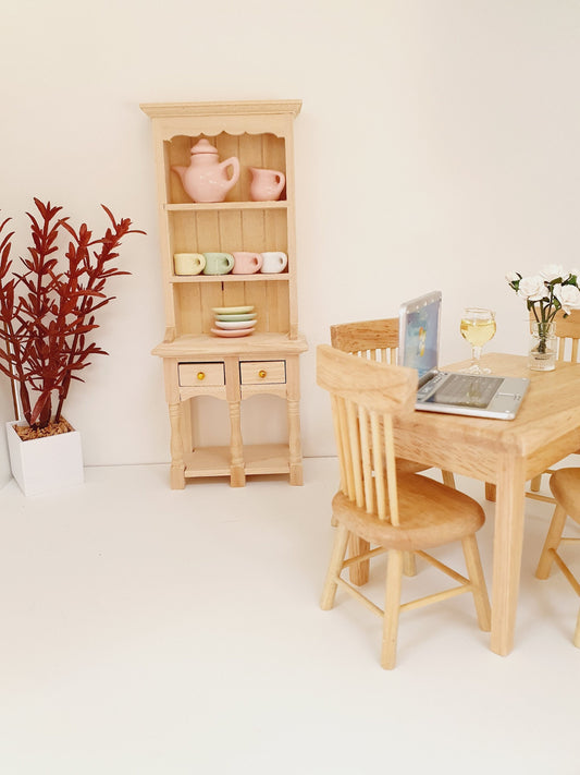 May include: A miniature wooden hutch with two drawers and open shelves, displaying a pink teapot, a white creamer, and three pastel-coloured teacups. A wooden table with two chairs and a laptop computer is in the foreground.