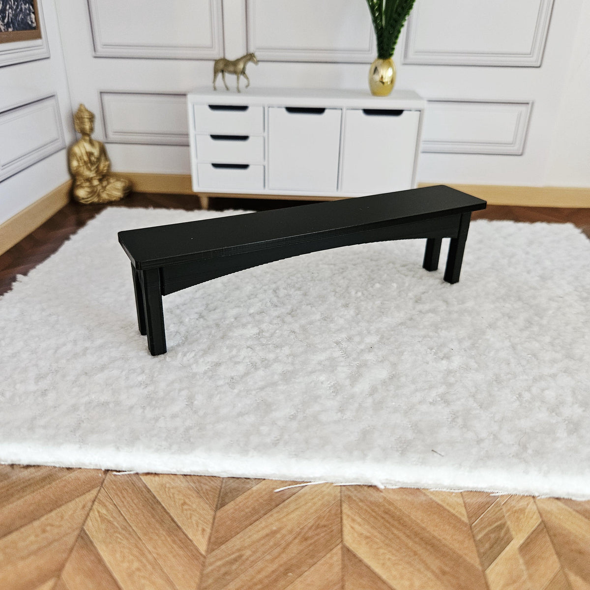 May include: A miniature black bench with a curved seat and angled legs. The bench is placed on a white, fluffy rug. The background includes a white cabinet, a gold statue, and a golden Buddha statue.