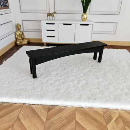 May include: A miniature black bench with a curved seat and angled legs. The bench is placed on a white, fluffy rug. The background includes a white cabinet, a gold statue, and a golden Buddha statue.