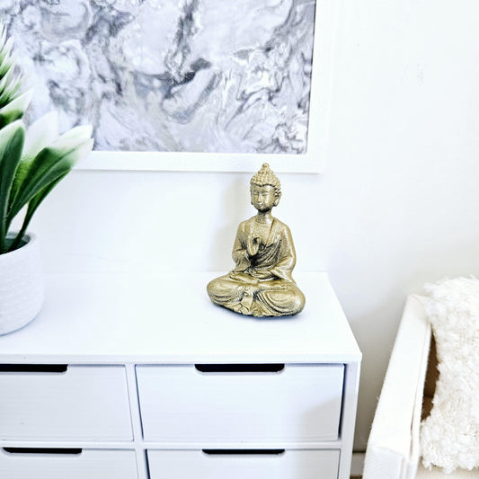 May include: A gold-coloured Buddha statue in a seated meditation pose. The statue is on a white chest of drawers. A framed abstract art piece hangs above the chest of drawers. A potted plant is on the left side of the image.