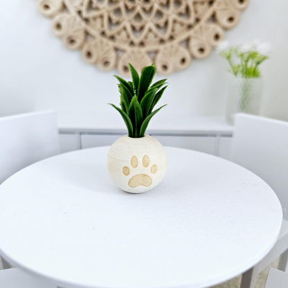 May include: A small white ceramic planter with a paw print design, holding a green artificial plant. The planter is on a white table.