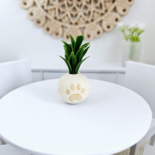 May include: A small white ceramic planter with a paw print design, holding a green artificial plant. The planter is on a white table.
