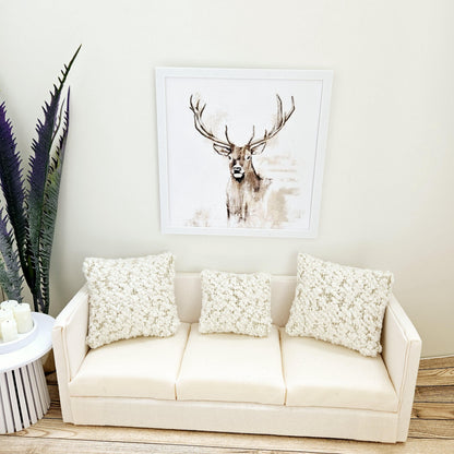 May include: A white sofa with three white cushions. The sofa is in front of a white wall with a framed print of a brown deer with large antlers.