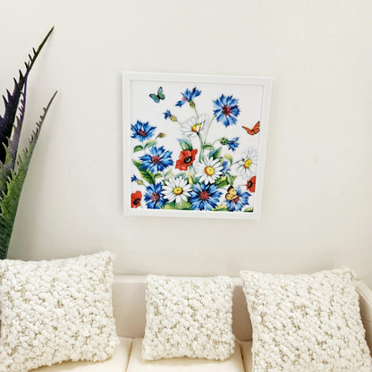 May include: A white framed picture with a floral print of blue, white, and red flowers with butterflies. The picture is hanging on a white wall above a white sofa with three white cushions.