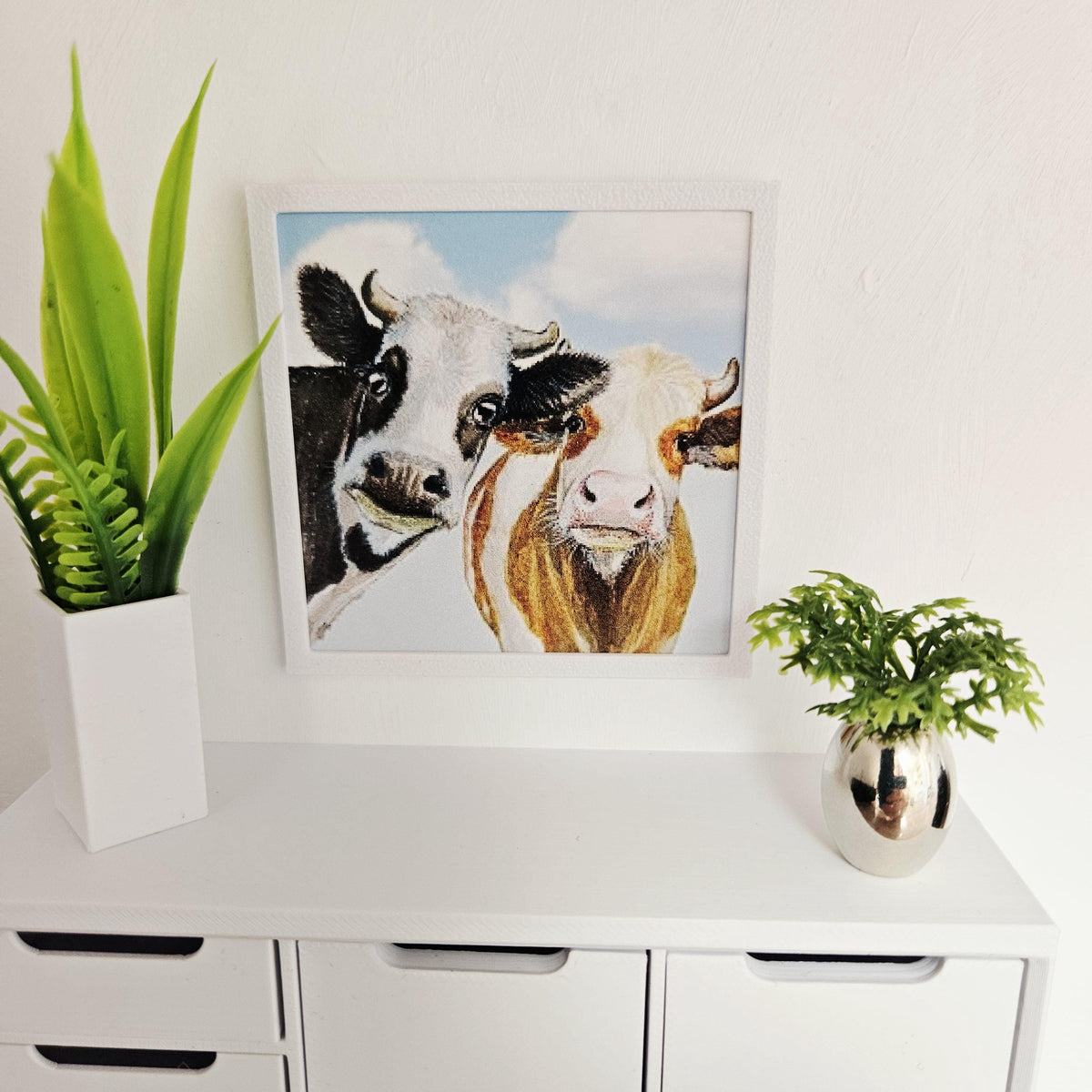 May include: A framed watercolour painting of two cows, one black and white, the other brown and white, against a blue sky with clouds. The artwork is displayed on a white chest of drawers with a green plant in a white vase and a small silver vase.