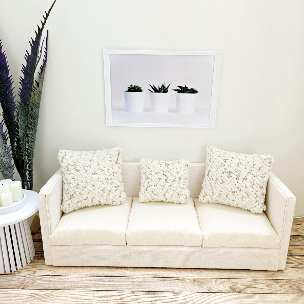 May include: A white sofa with three white throw cushions. The sofa is in front of a white wall with a framed print of three potted succulents.