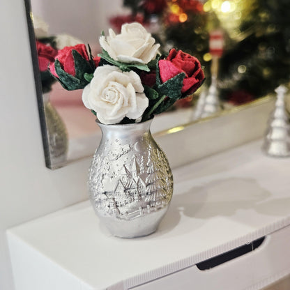 Red and white roses in a silver vase with raised village decoration, standing on a white sideboard in front of a mirror.