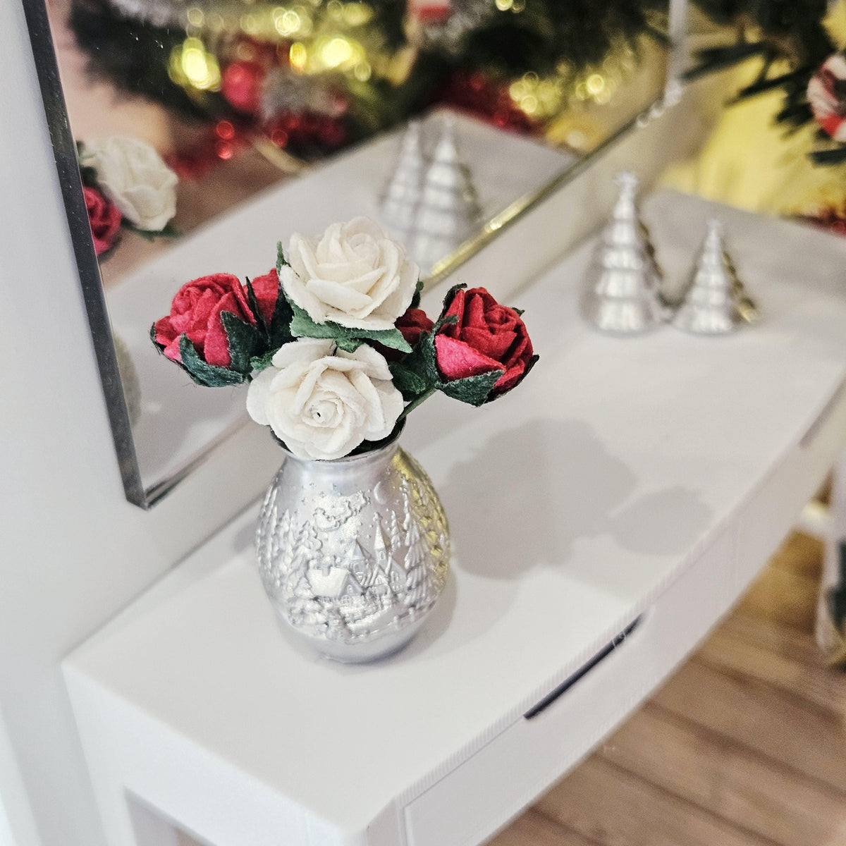 Elevated view of red and white roses in a silver Christmas vase on a white sideboard, with a mirror and silver tree decorations behind.