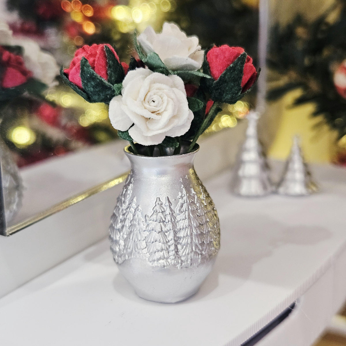 Silver Christmas vase showing raised fir tree decoration beneath red and white roses, with miniature silver trees in the background.