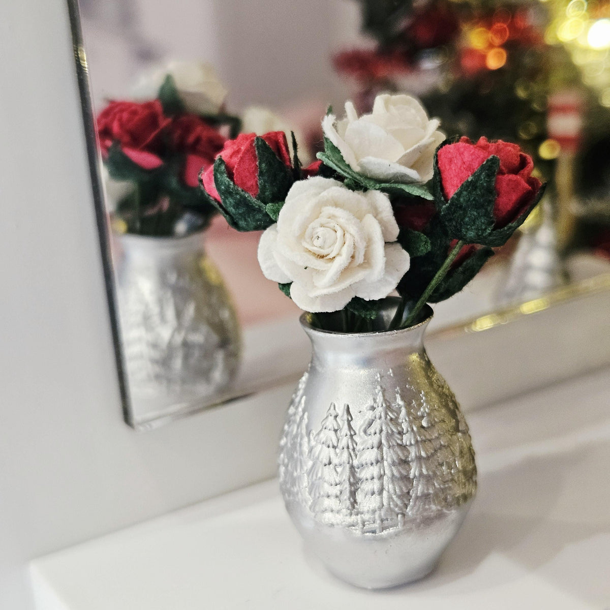 Close view of red and white roses with green foliage in a silver vase, showing raised fir trees and its reflection in a mirror.