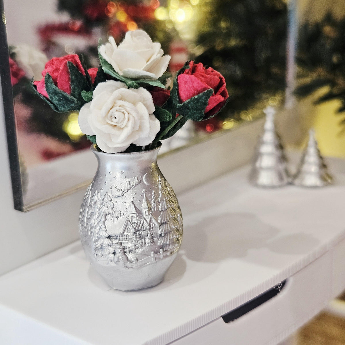 Silver Christmas vase with red and white roses, green foliage and a raised village design, displayed on a white sideboard beside a mirror.