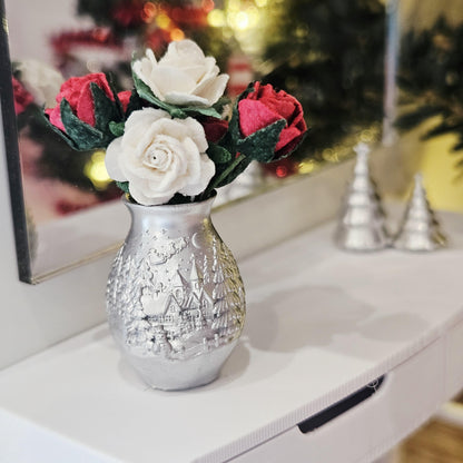 Silver Christmas vase with red and white roses, green foliage and a raised village design, displayed on a white sideboard beside a mirror.