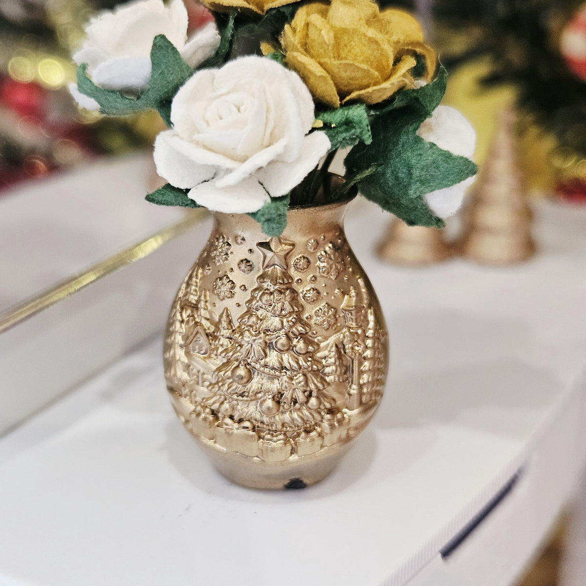 Close-up of the raised Christmas tree decoration on the gold-coloured vase, beneath white and gold roses and green leaves.