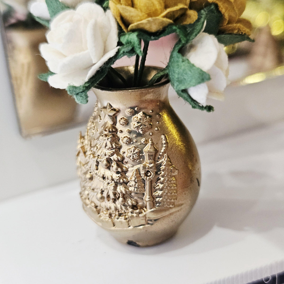 Side close-up of the gold-coloured vase showing raised festive decoration, its open neck and rose stems beneath green leaves.