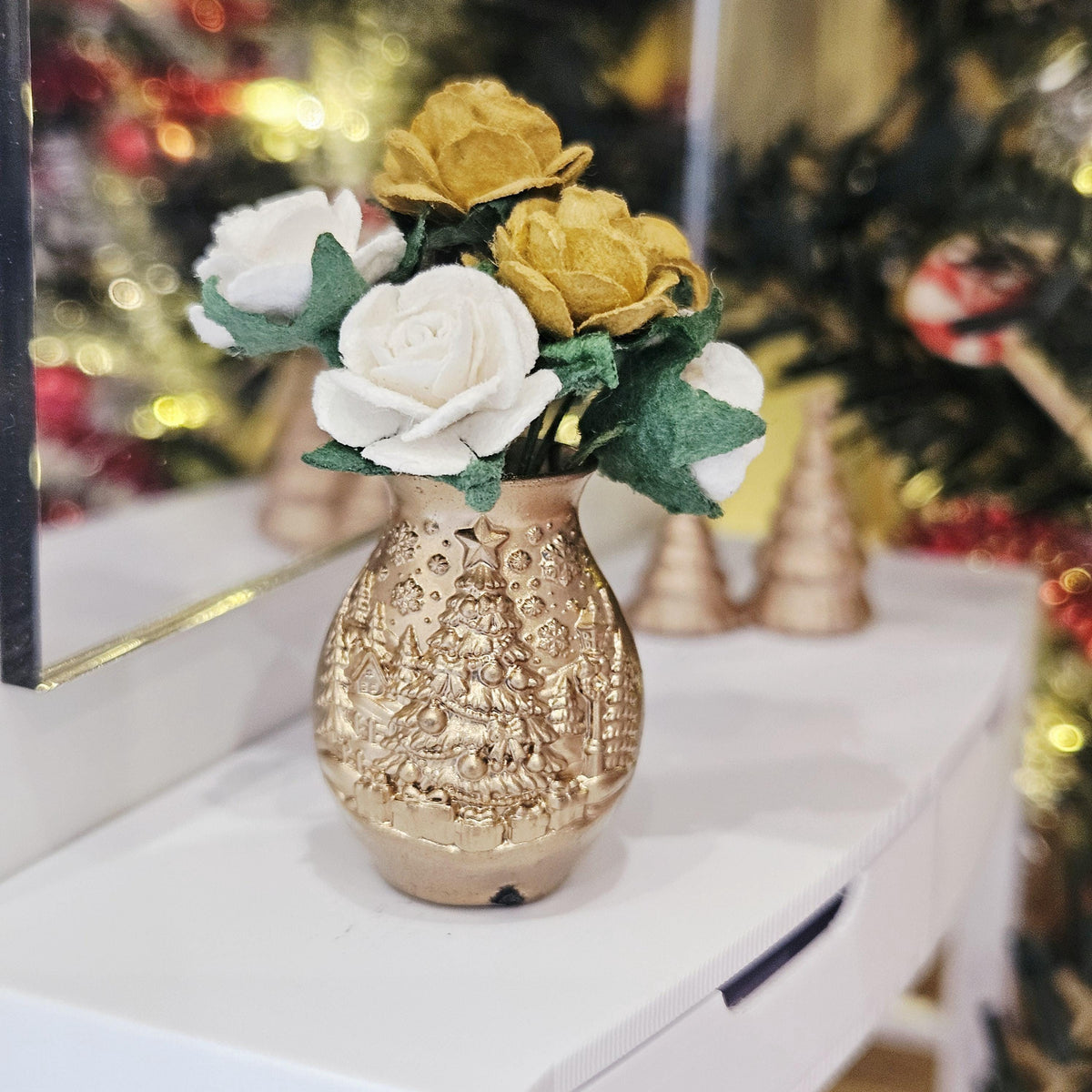 Gold and white roses in a gold-coloured Christmas tree vase on a white sideboard, with a decorated Christmas tree behind.