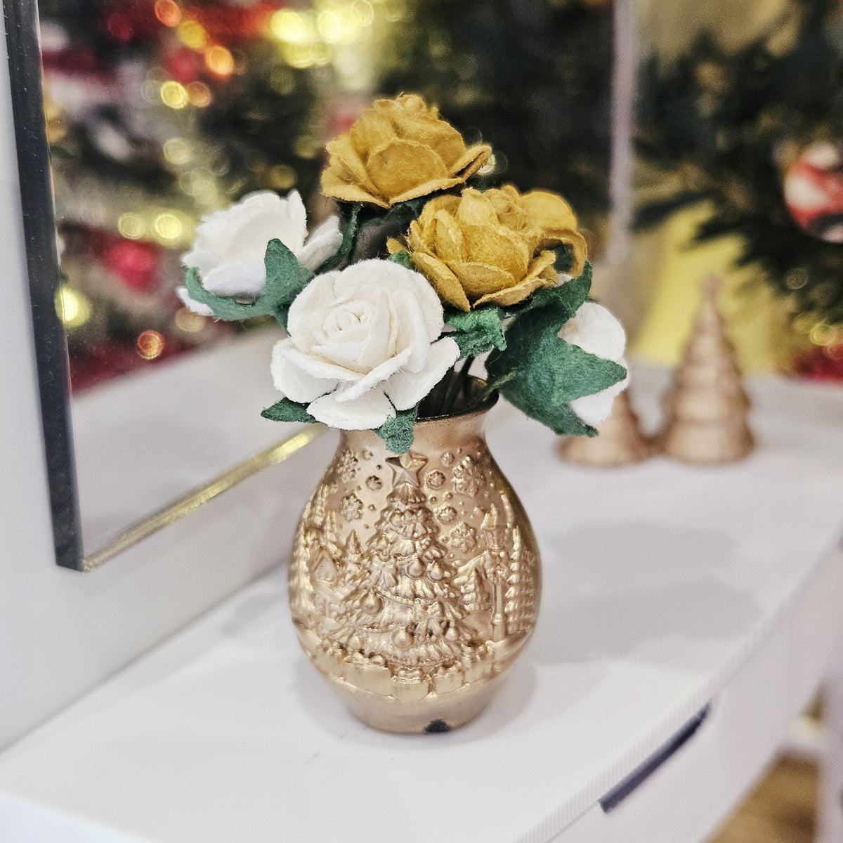 Gold-coloured Christmas tree vase holding gold and white roses with green leaves, displayed on a white sideboard beside a mirror.