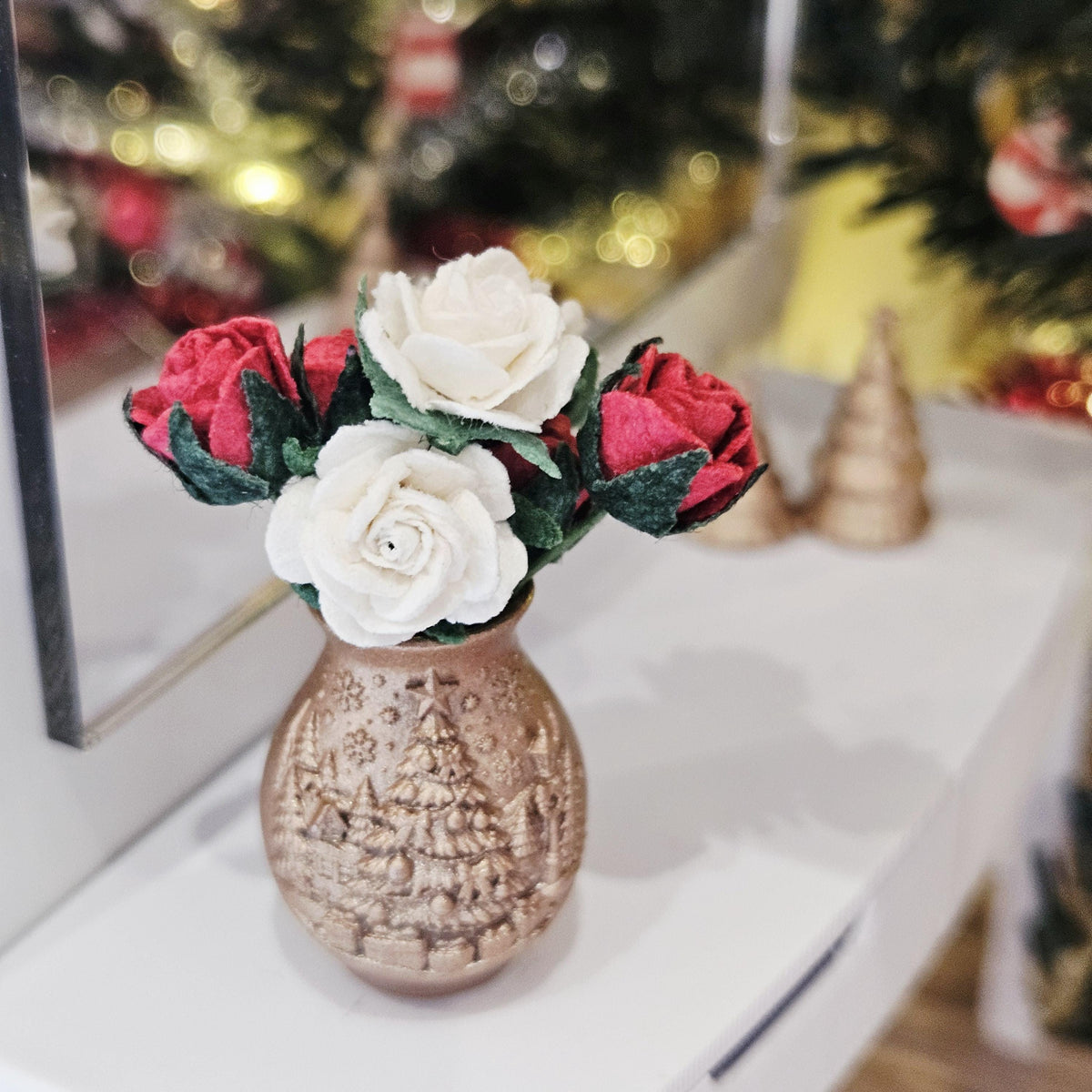 Red and white roses with green leaves in a copper-coloured Christmas tree vase, standing on a white sideboard beside a mirror.
