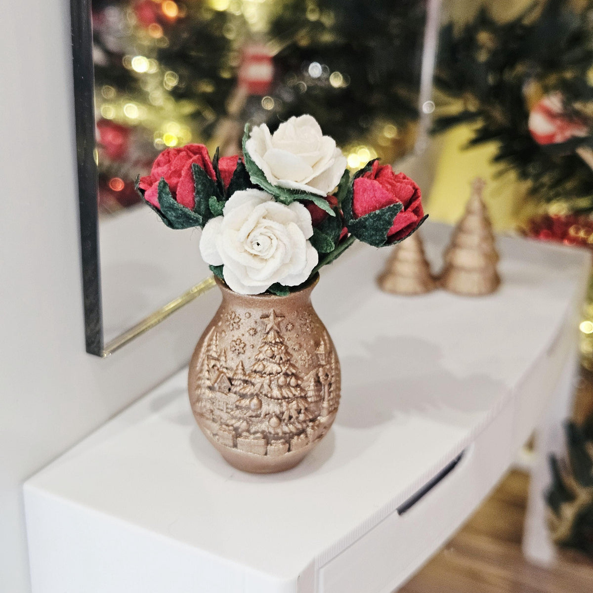 Front view of the copper-coloured vase with raised Christmas tree decoration and red and white roses, with miniature trees behind.