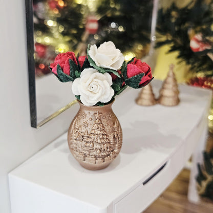 Front view of the copper-coloured vase with raised Christmas tree decoration and red and white roses, with miniature trees behind.