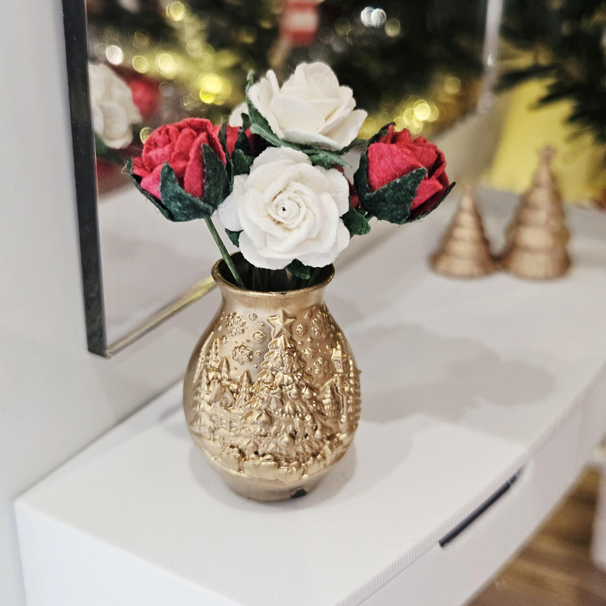 Red and white roses in a gold-coloured vase with raised Christmas tree decoration, displayed on a white sideboard near a mirror.